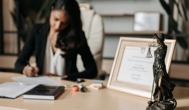 Focused woman writing at desk with Lady Justice figurine and certificate in office.